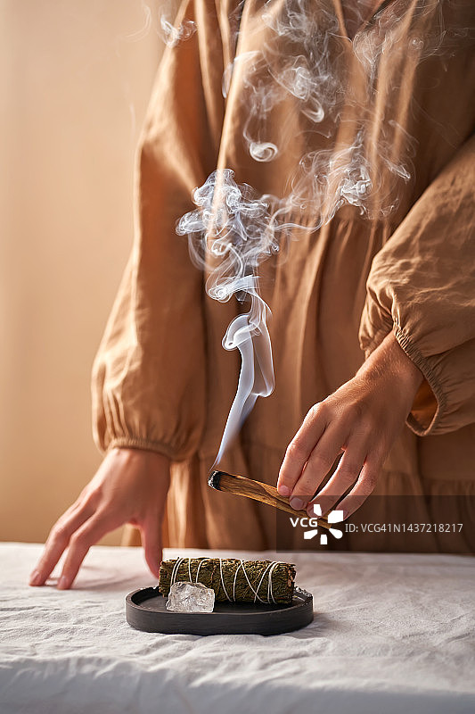 一个穿着米色衣服的女孩的手拿着一根冒烟的帕罗桑托木棍，背景是仪式上的东西:一块水晶石和一根用来熏蒸的麻花。图片素材