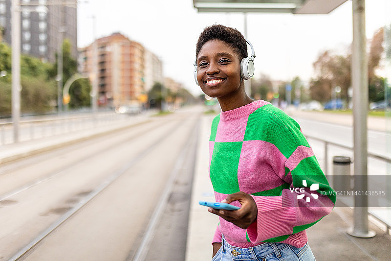微笑漂亮的年轻成年非洲时髦女子拿着手机，戴着耳机听音乐站在电车车站图片素材