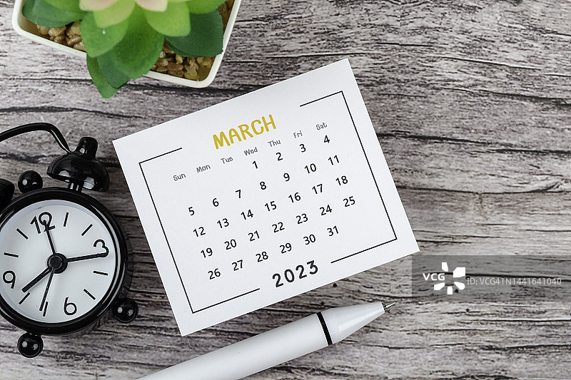 日历桌2023:3月是组织者计划和截止日期的月份，木桌背景上有一个闹钟和白笔。图片素材