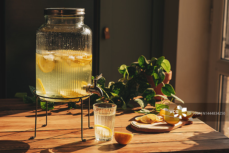 柠檬水加冰装在水罐里，玻璃杯放在木桌上，外面有水果和碎冰，背景是一片柠檬田，天气晴朗。前视图。水平组合。图片素材