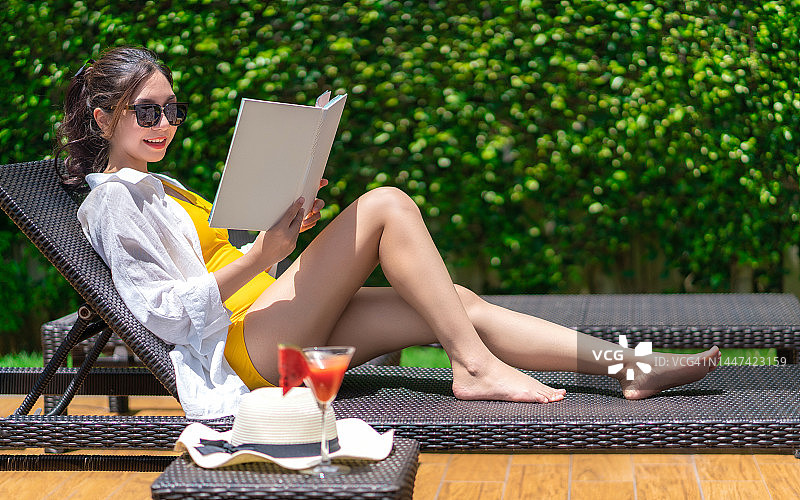 年轻美丽的亚洲女人戴着太阳镜躺在躺椅上享受日光浴日光浴泳装阅读书在游泳池边暑假。女人阅读和放松附近的豪华游泳池图片素材