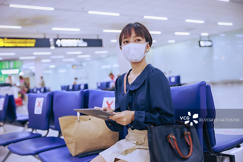 年轻的亚洲女商人戴着口罩在机场离境区用平板电脑阅读电子邮件。商务旅行的概念。图片素材