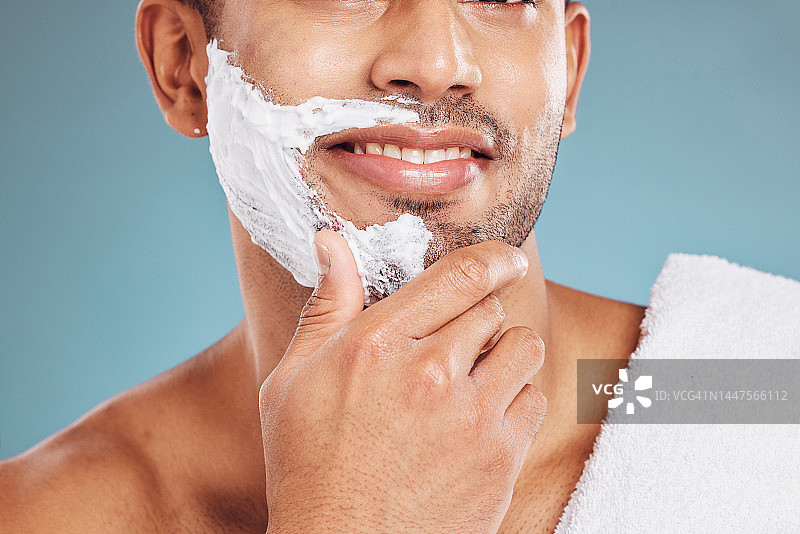 男子与剃须膏在脸上，胡须保养和护肤，男性健康和美容治疗与蓝色背景。健康，皮肤护理和面部毛发，早上用毛巾剃须。图片素材
