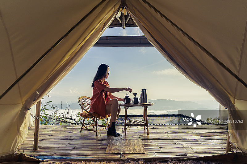 早晨，一个女人坐在自己的帐篷前喝咖啡，看着雾蒙蒙的大海。阳光从右边照进来。图片素材