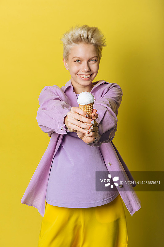 专注于冰淇淋。在黄色背景的摄影棚里，年轻微笑的少女手里拿着冰淇淋甜筒图片素材