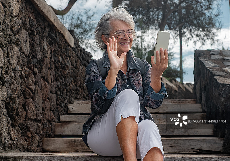 英俊的高级女子坐在户外的木楼梯上，一边用手机视频聊天。快乐微笑的老妇人使用无线技术图片素材