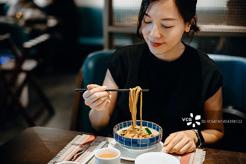 在一家中国餐馆里，年轻漂亮的亚洲女子正在用筷子吃着一碗面，喝着一杯茶图片素材