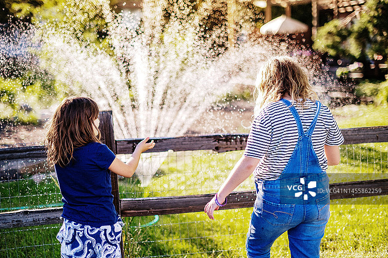两个女孩在户外玩着院子里的洒水器和尖桩篱笆图片素材