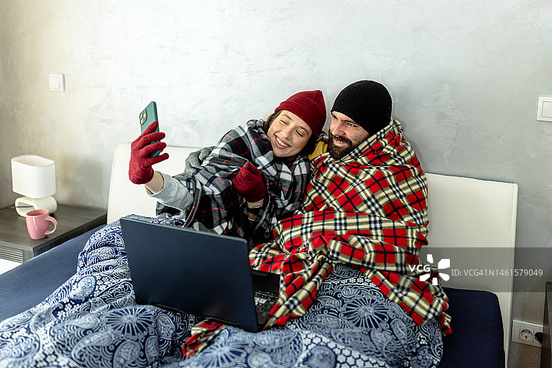一对穿着暖和衣服的夫妇在家里冻得发抖，在卧室里盖着毯子试图暖和起来。图片素材