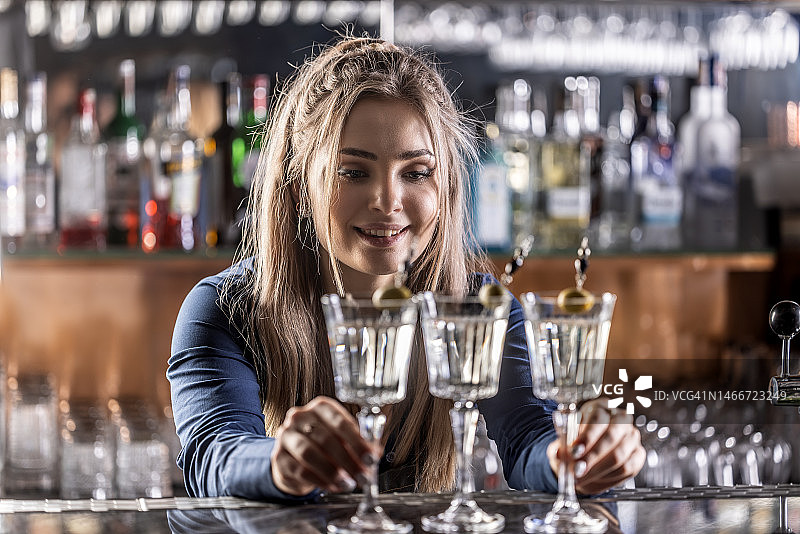 年轻的酒吧女服务员在吧台上准备了三杯干马提尼鸡尾酒。图片素材