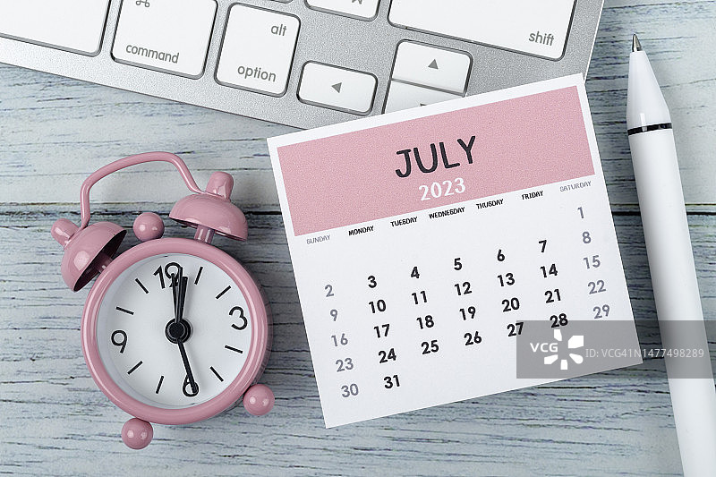 日历桌2023:7月是组织者计划和截止日期的月份，木制桌子背景上有闹钟和电脑键盘。图片素材