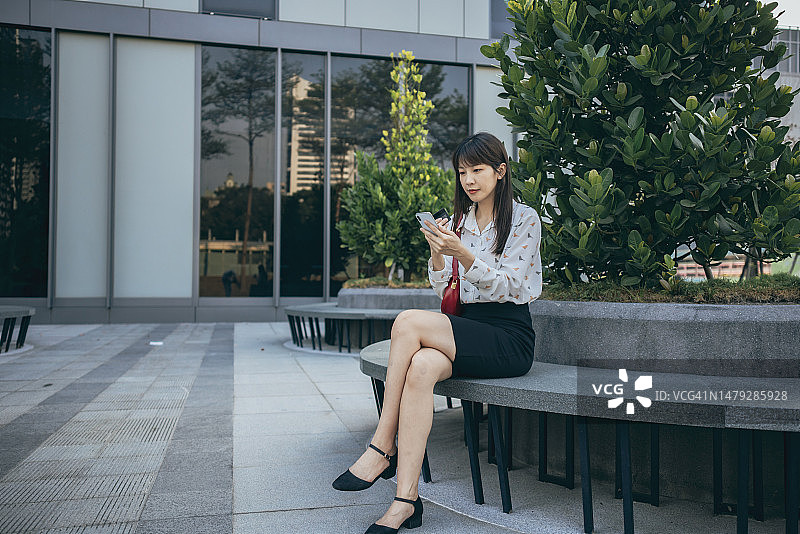 一位年轻的亚洲女子坐在城市公园里，用她的智能手机在网上购物，手里拿着一张信用卡进行移动支付。这个场景说明了网上购物和移动支付的概念。图片素材