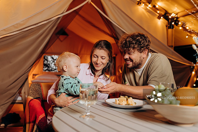 幸福的家庭和可爱的宝宝一起吃晚餐，在夏天的晚上在舒适的篝火旁度过美好的时光。豪华露营帐篷，适合户外休闲娱乐。生活方式的概念图片素材