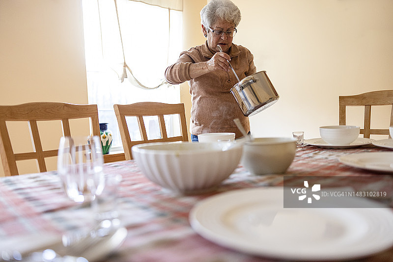 一位白发苍苍的老妇人用勺子把锅里的汤舀进碗里，为家人提供晚餐。图片素材