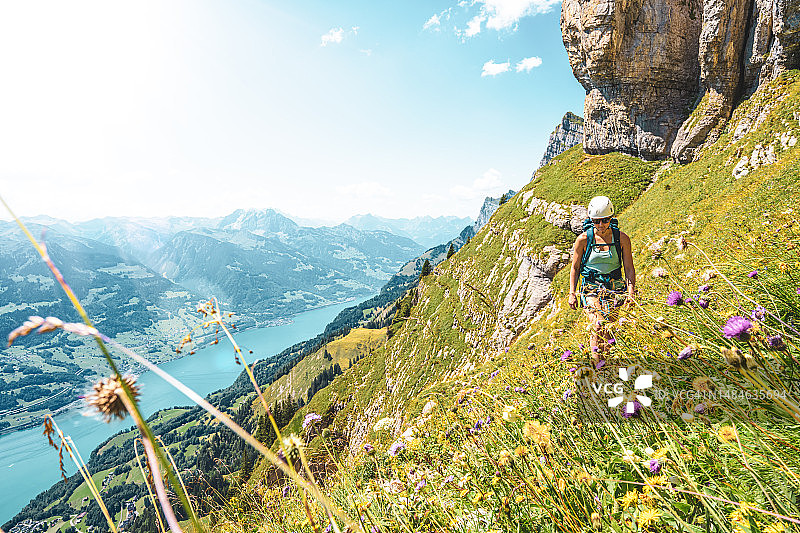 运动女子徒步旅行的花草地旁边陡峭的岩石墙与风景秀丽的瓦伦泽和瑞士阿尔卑斯山的背景。瑞士，瓦伦塞，圣加仑，瑞士，欧洲。图片素材
