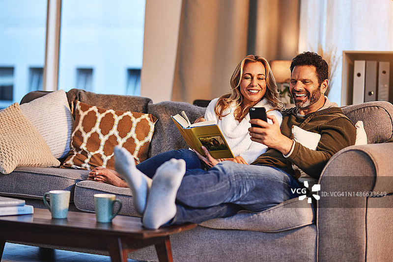 幸福的夫妻，在家里的沙发上打电话和笑，有互联网连接和健康的婚姻。成熟的女人读一本书，为一个拿着智能手机的男人笑，看一个有趣的表情包或视频，享受美好时光图片素材