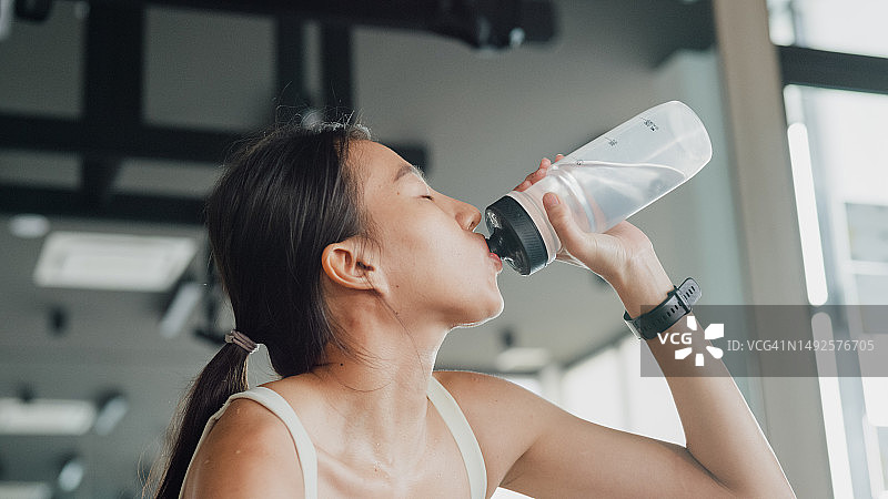 现代健身房里，年轻的亚洲女运动员在运动，健美运动员在喝瓶装水。图片素材