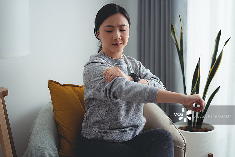 一位亚洲妇女坐在家里客厅的扶手椅上，忍受着手臂疼痛。图片素材
