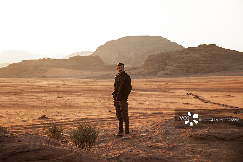 一个年轻人双手插在口袋里，站在日落时分的沙漠里图片素材