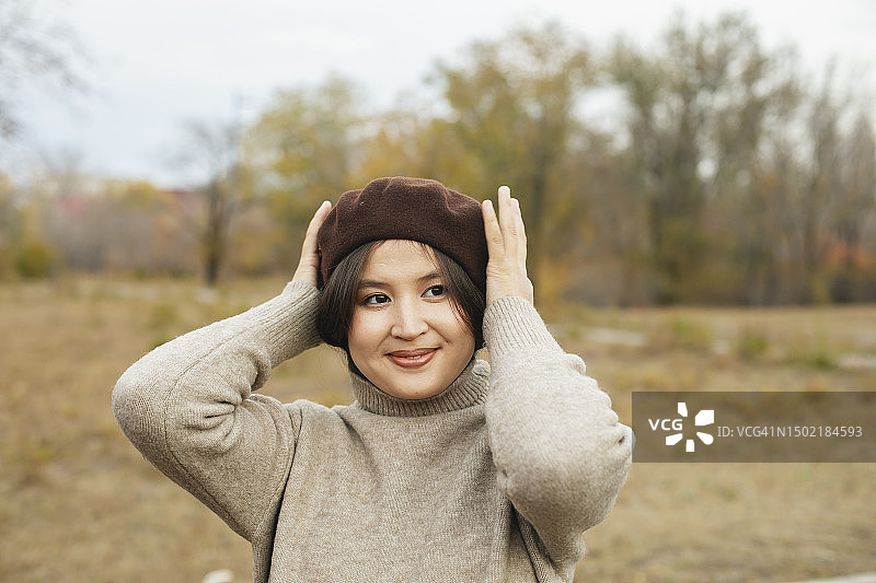 在秋天的户外，穿着米色毛衣和棕色贝雷帽的年轻亚洲女人微笑的肖像图片素材