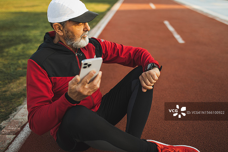 一位老人坐在运动跑道上休息。将智能手表与手机应用程序同步，查看跑步结果图片素材