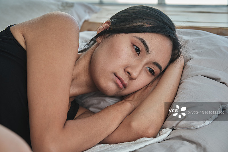 悲伤的亚洲女性躺在床上望着远方，感到孤独、悲伤，正在应对离婚或分手。忧郁的日本千禧一代女孩在家。精神痛苦概念图片素材