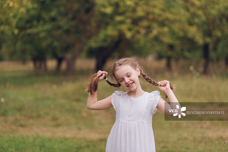 一个快乐的小女孩带着辫子走在一个苹果园带着微笑的肖像-库存照片图片素材
