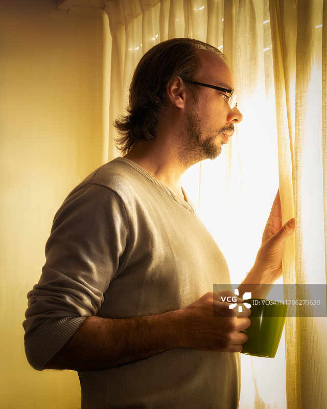 一名男子拿着咖啡杯，透过窗户的窗帘往外看图片素材