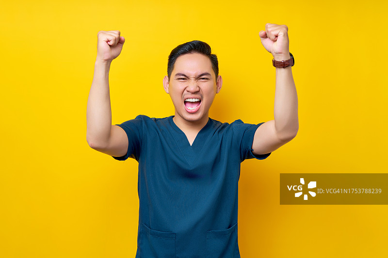 兴奋的年轻专业亚洲男医生或护士穿着蓝色制服，自信地站在黄色背景上庆祝成功。保健医学概念图片素材