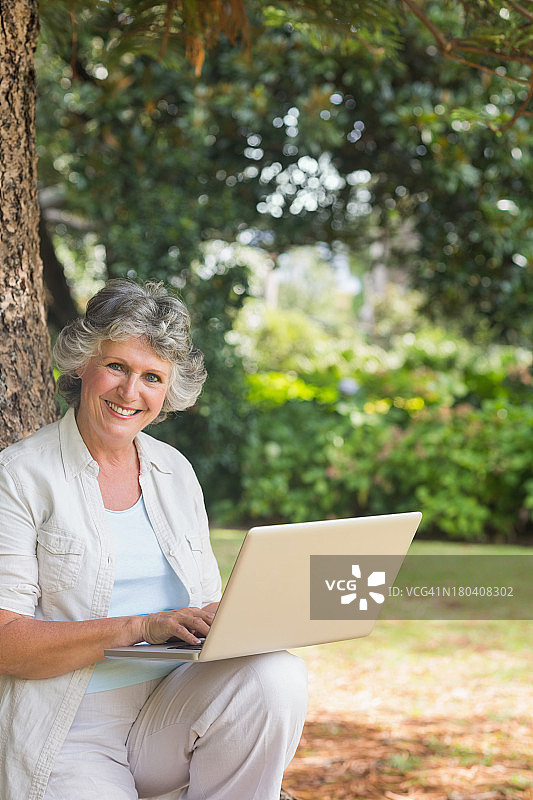 快乐成熟的女人坐在树干上用笔记本电脑图片素材