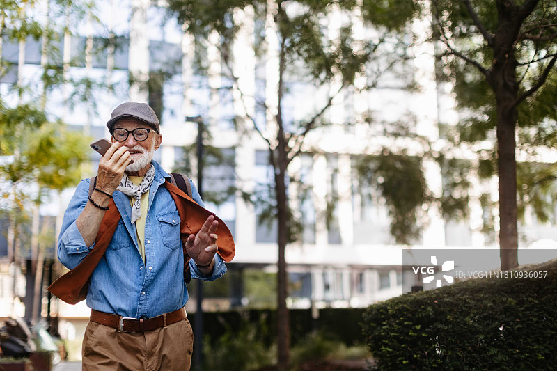 一位城市老人拿着智能手机，拿着免提打电话。老人走在城市街道上，与家人通话。图片素材