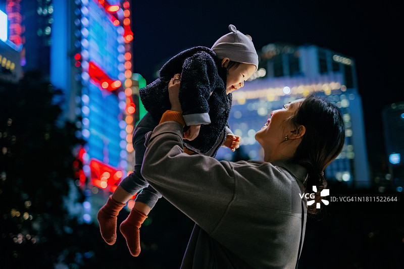 微笑的年轻亚洲母亲在空中抱着她的女儿，站在城市的夜晚，面对着灯火通明、色彩缤纷的城市摩天大楼。规划未来。有孩子的家庭。希望与期待概念图片素材