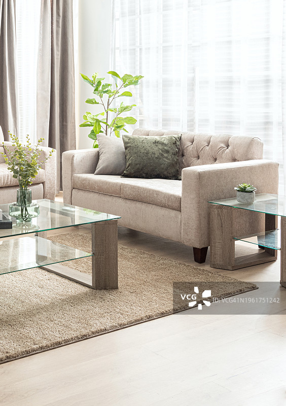 客厅内饰米色面料沙发套和矩形玻璃咖啡桌与架子和木腿。房间有一扇窗户，米色亚麻窗帘，淡绿色墙壁。图片素材