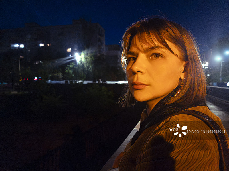 平静美丽的成年女子，站在夜晚的城市街道上。自信的看图片素材