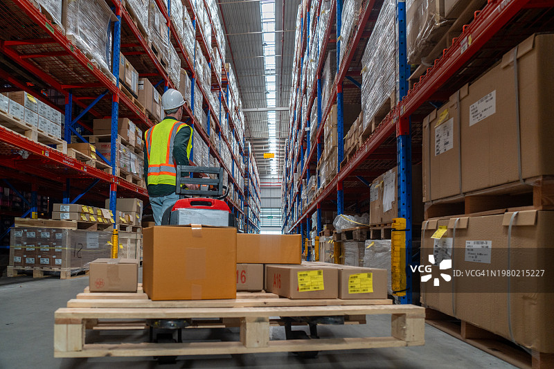 零售配送仓库的货架上堆满了纸箱里的货物，工人扫描和分类包裹，用托盘卡车移动库存。图片素材