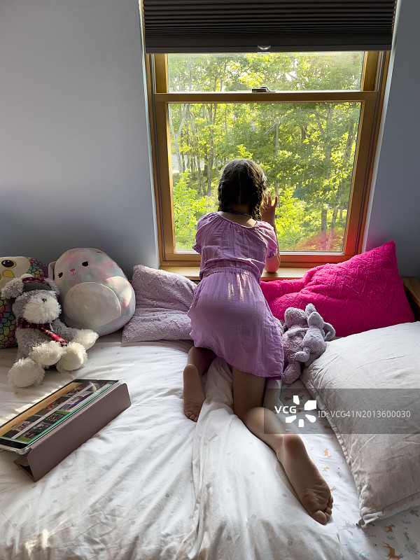 6岁的小女孩从后面看卧室窗外，她把iPad放在床上，手放在窗户上图片素材