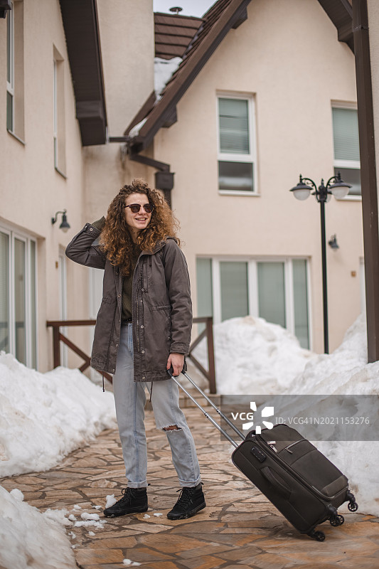 一位年轻的女旅行者提着行李箱站在一家旅馆门前，她是来度寒假的图片素材