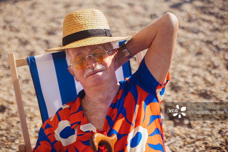 老人闭着眼睛躺在沙滩椅上打盹的照片。老人在海边放松，享受暑假。退休后旅行的概念。图片素材