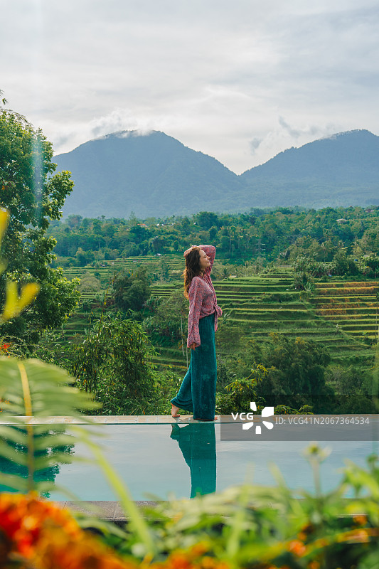优雅的女子站在泳池边，俯瞰着巴厘岛的稻田图片素材