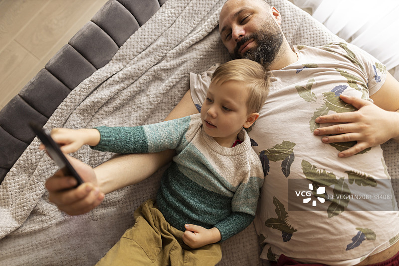 爸爸和小儿子笑着看着手机上的照片。他们躺在卧室的床上拍照。父亲拥抱儿子。他们做鬼脸。一个深色头发和浓密胡须的男人和一个浅色头发和眼睛的男孩图片素材