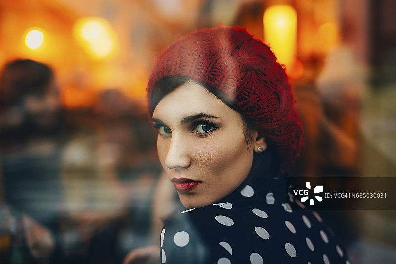 一个年轻女子戴着红帽子在晚上透过窗户看酒吧的肖像图片素材
