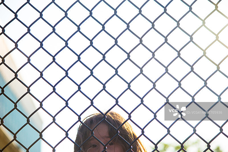 一个小女孩透过铁链栅栏往外看。图片素材