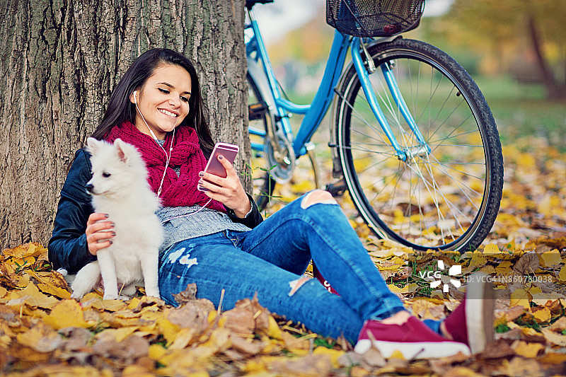 美丽的女孩坐在秋天的树叶上，和她的小狗一起听音乐图片素材