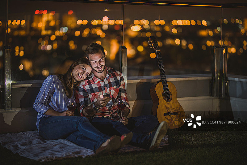 一对年轻夫妇晚上在顶层公寓的露台上使用智能手机。图片素材