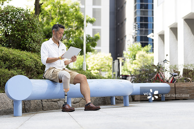 一名日本男子戴着假肢坐在办公室外，手里拿着一个数字平板电脑图片素材