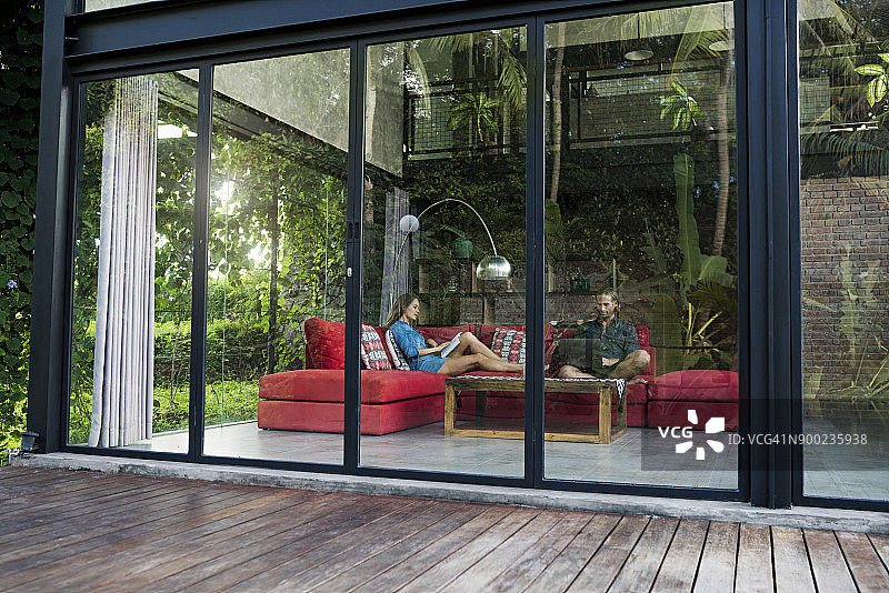在现代设计的房子里，一对夫妇带着笔记本电脑和书籍放松在红色的沙发上，透过玻璃立面的花园景观图片素材