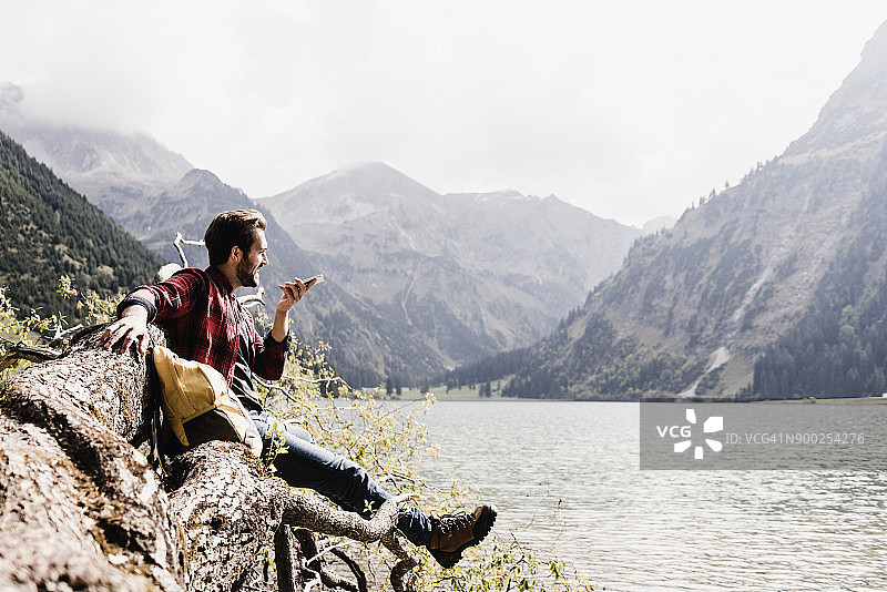 奥地利，蒂罗尔，阿尔卑斯山，徒步旅行者在山上的湖泊的树干上使用手机图片素材