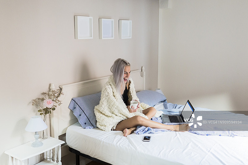 快乐的年轻女人躺在床上，拿着咖啡和笔记本电脑图片素材