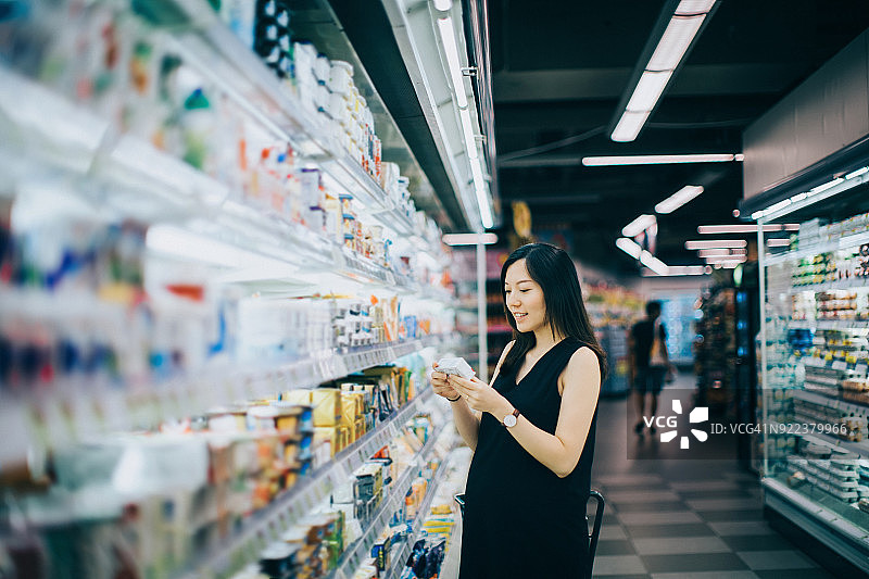 亚洲孕妇在超市购物，阅读一包奶酪上的营养标签图片素材