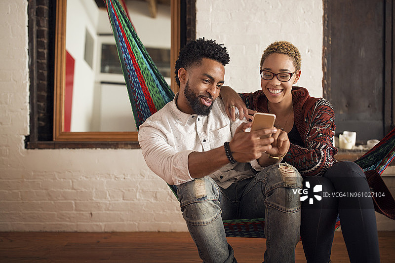 一对快乐的年轻夫妇坐在家里的吊床上玩智能手机图片素材
